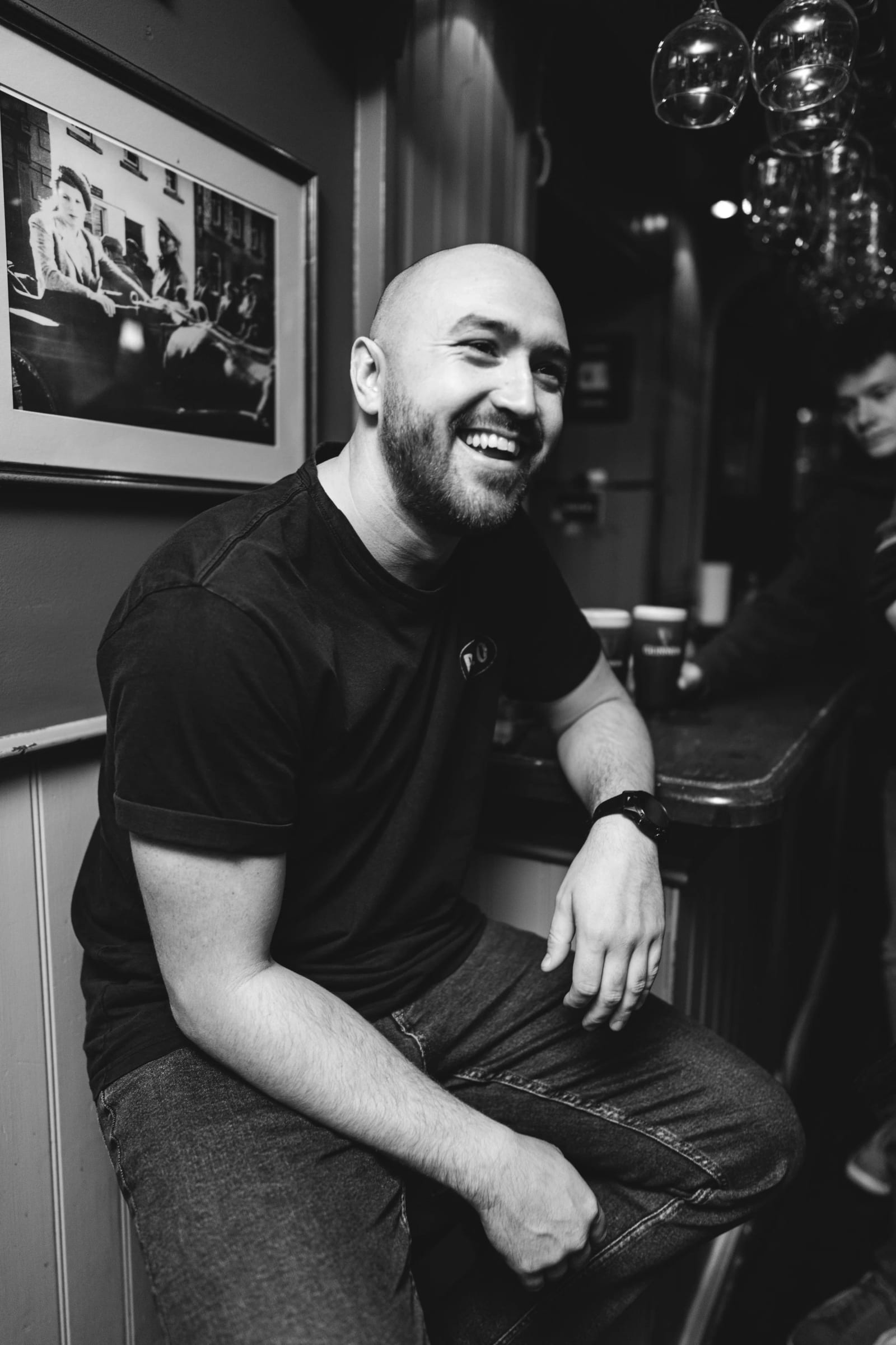 Black and white photograph of Kieran J Edwards laughing in a black T-shirt, leaning against a bar beneath framed pictures.