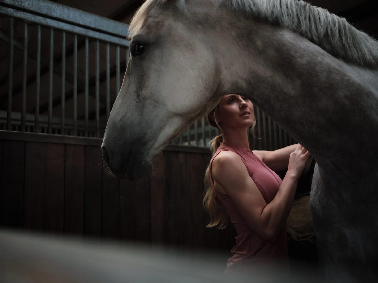 A woman in a pink top standing close against a grey horse inside a stable.