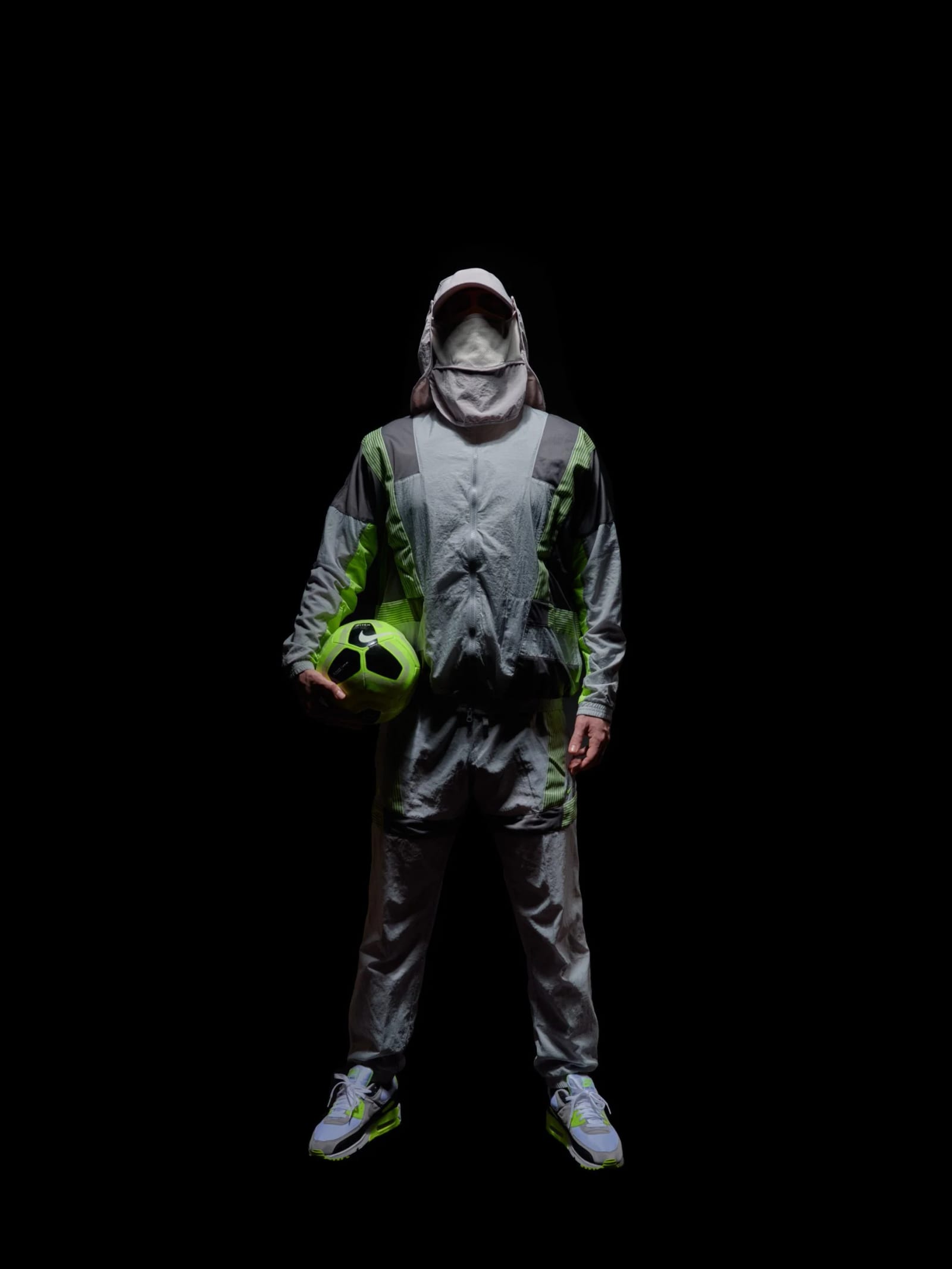 A figure in a hooded grey and green tracksuit with the face covered, holding a green football against a black background.