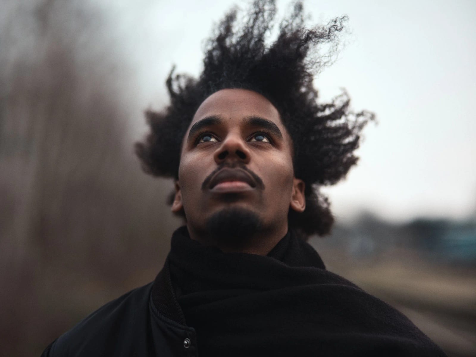 A man in a black coat looking upward, his hair blown sideways by the wind against a stormy sky.