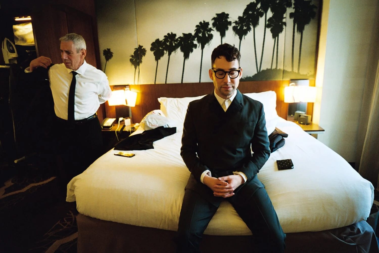 A man in a dark suit and glasses sitting on a hotel bed beneath a palm-tree mural, an older man in a shirt and tie standing beside it.