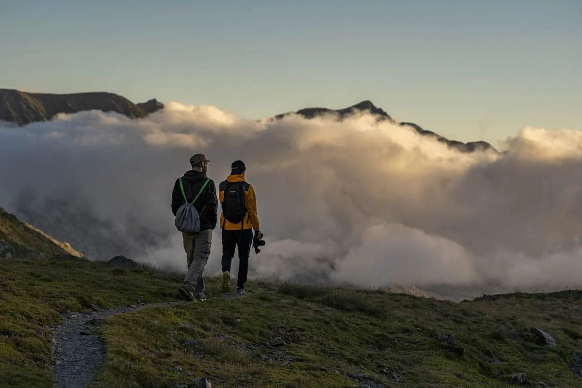 Two hikers with rucksacks walking a grassy ridge toward a bank of cloud at sunset.