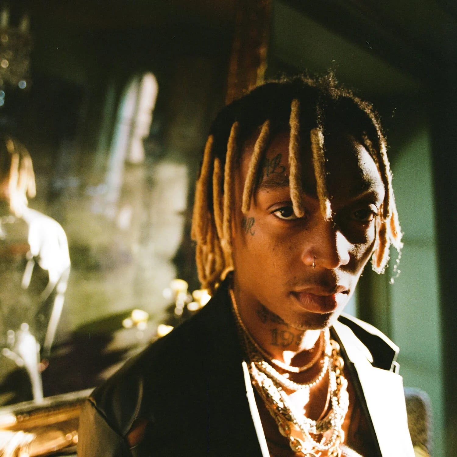 A young man with bleached twists and layered chains lit by warm side light, looking over his shoulder.