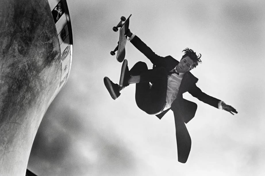 Black and white photograph of a man in a suit flying upside down mid-air with a skateboard above a ramp.