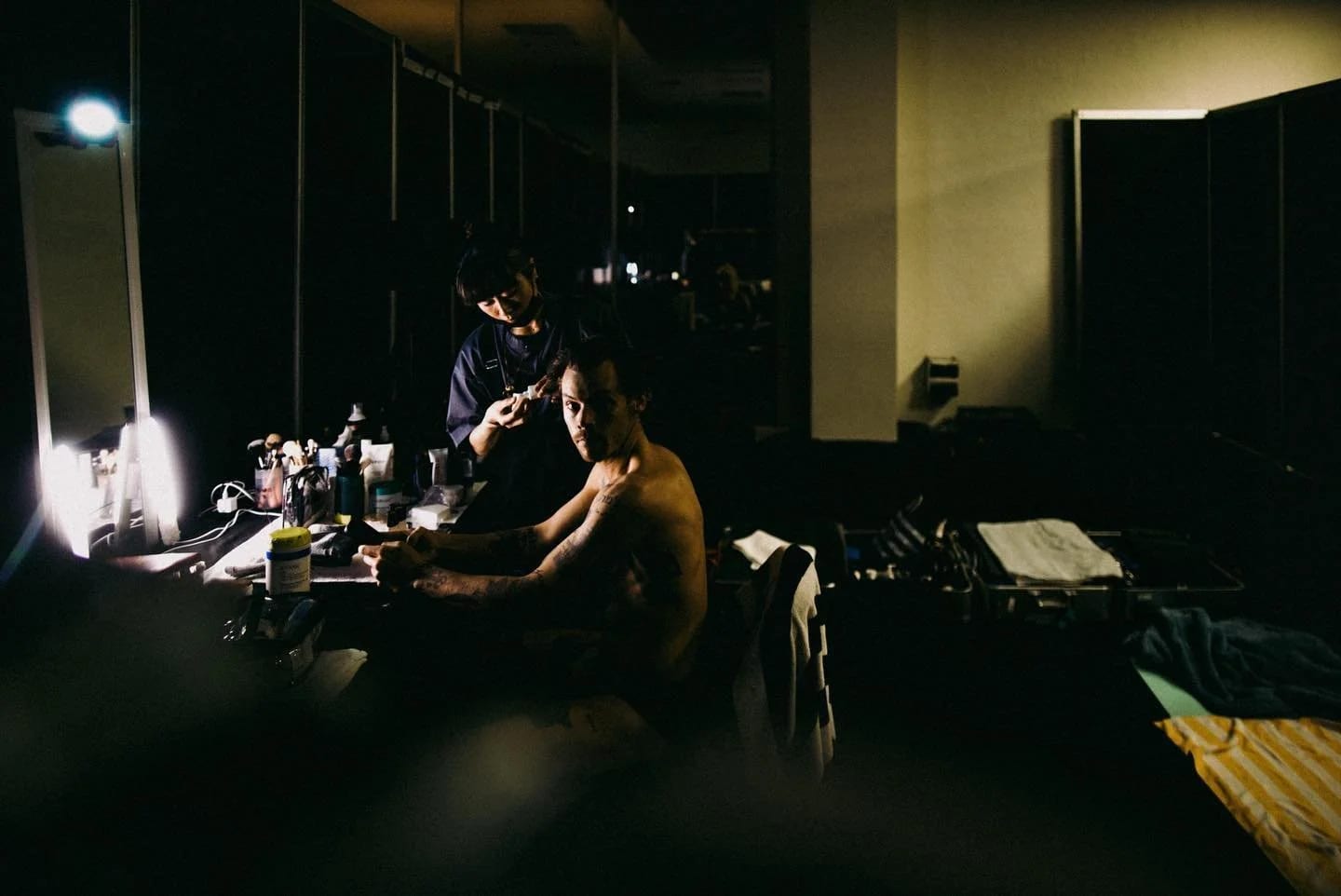 Harry Styles seated shirtless at a dressing-room mirror while someone works on his hair, the room dark around the lit mirror.