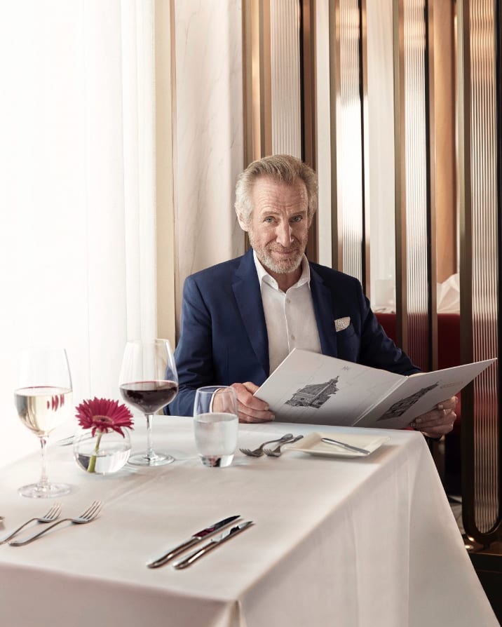 A grey-haired man in a blue blazer reading a menu at a restaurant table set with wine glasses.