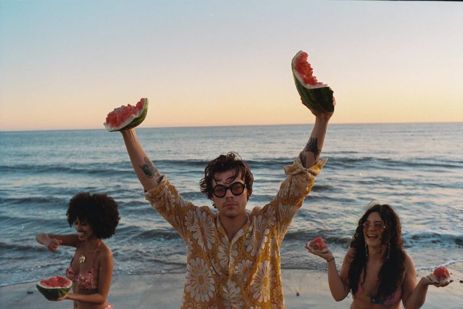 Harry Styles in a yellow floral shirt holding two slices of watermelon above his head at the shore, two women eating watermelon beside him.