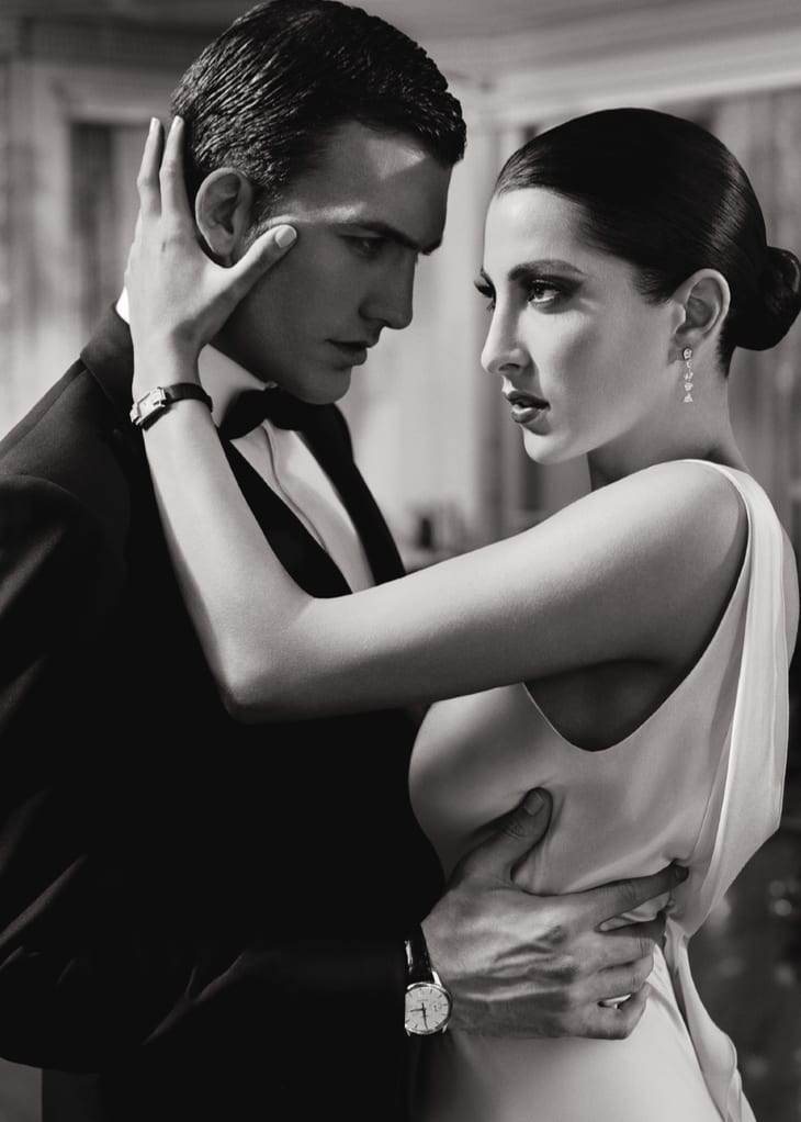 Black and white photograph of a man in a tuxedo and a woman in a white dress about to kiss, both wearing wristwatches.