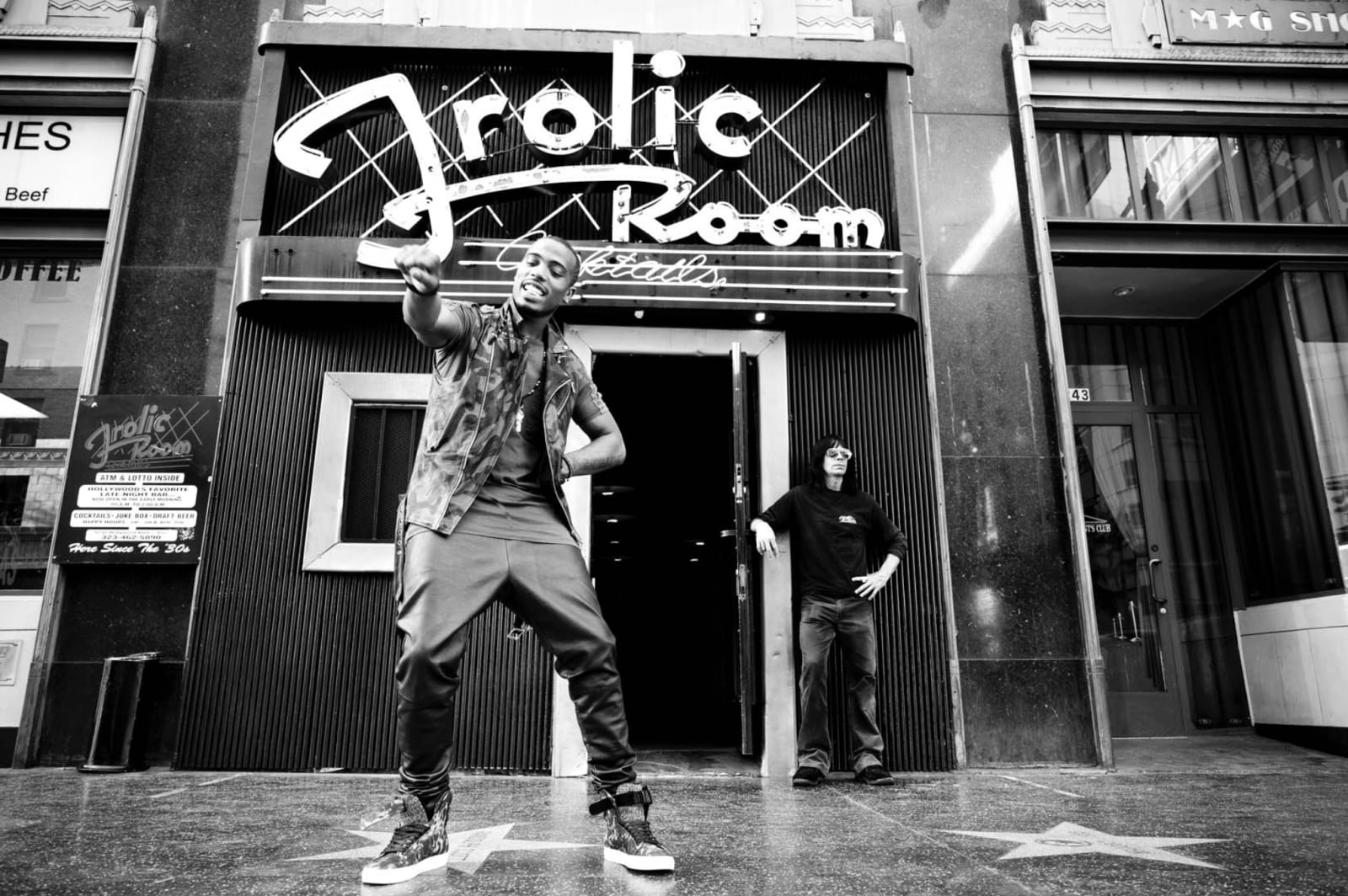 Black and white photograph of a man in a patterned shirt pointing at the camera outside a bar with a Frolic Room neon sign, stars on the pavement.