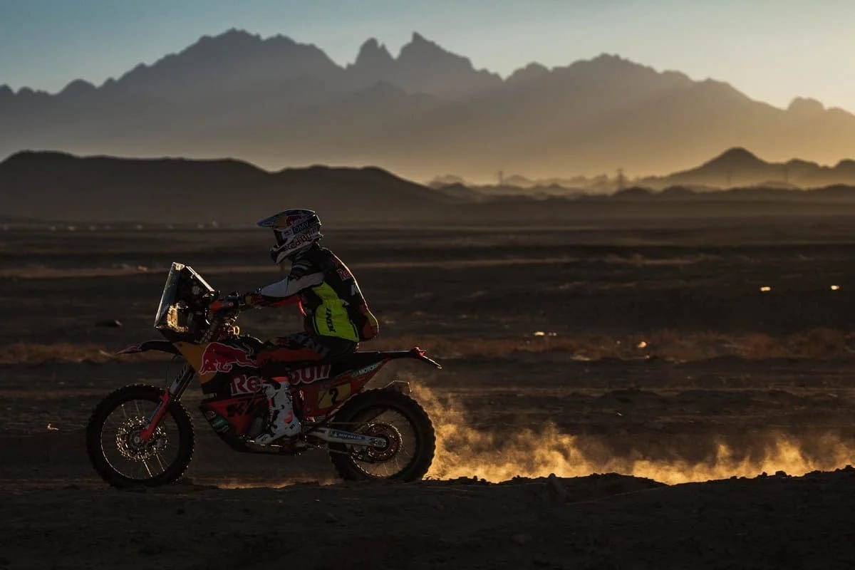 A rally motorcyclist crossing a dark desert plain at dawn, jagged mountains on the horizon.