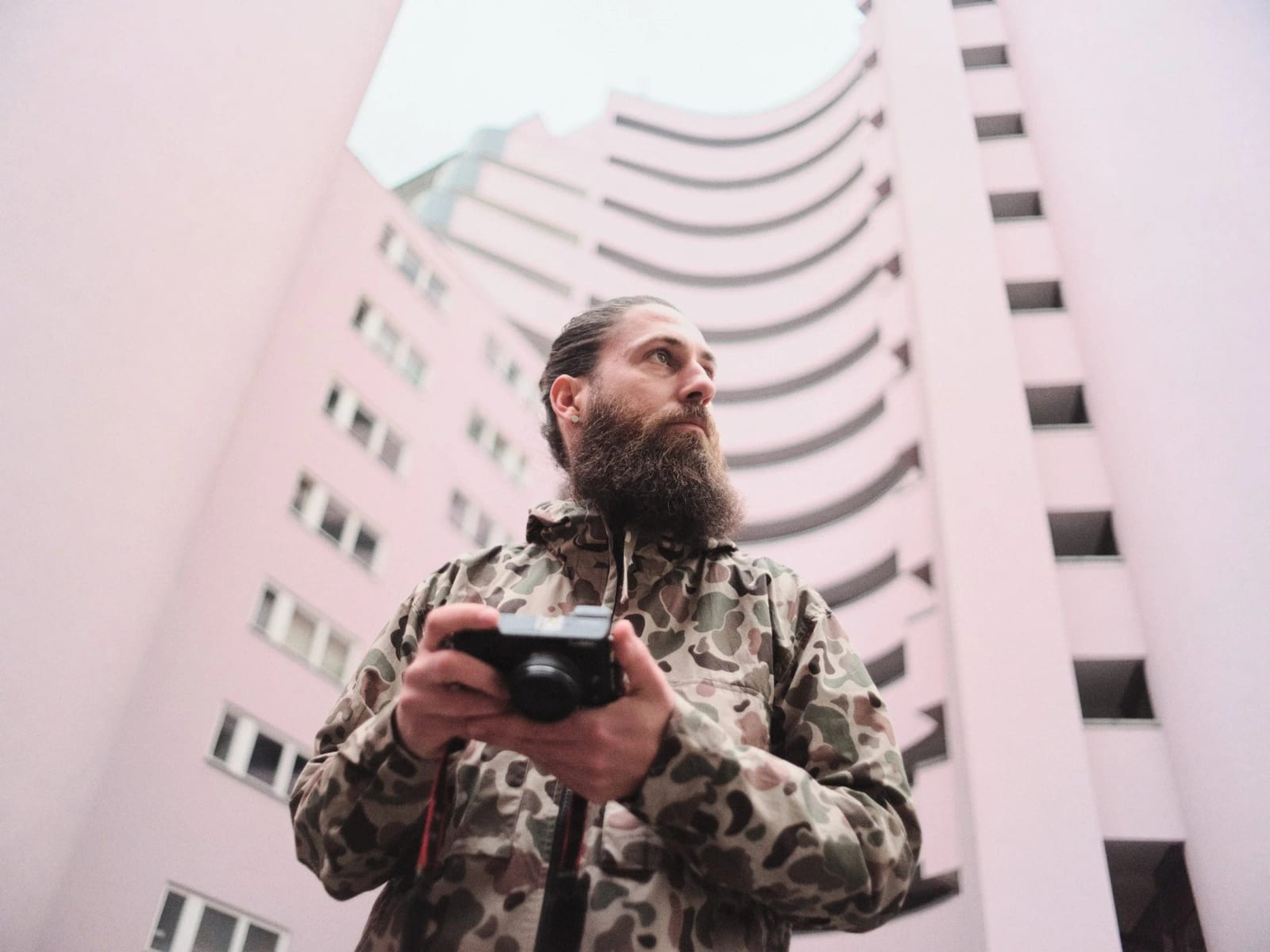 A bearded man in a camouflage jacket holding a camera, looking up beneath a pale pink tower block.