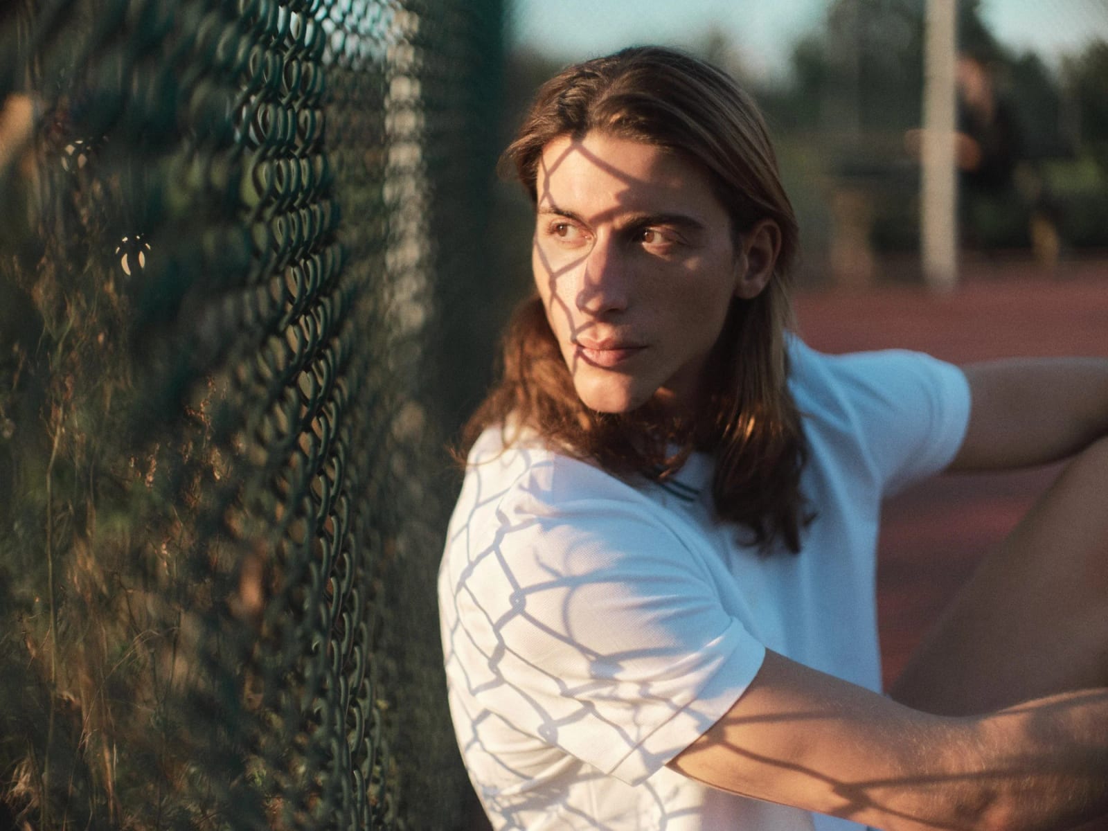 A long-haired man in a white T-shirt leaning against a chain-link fence, its shadow patterning his face and shirt.