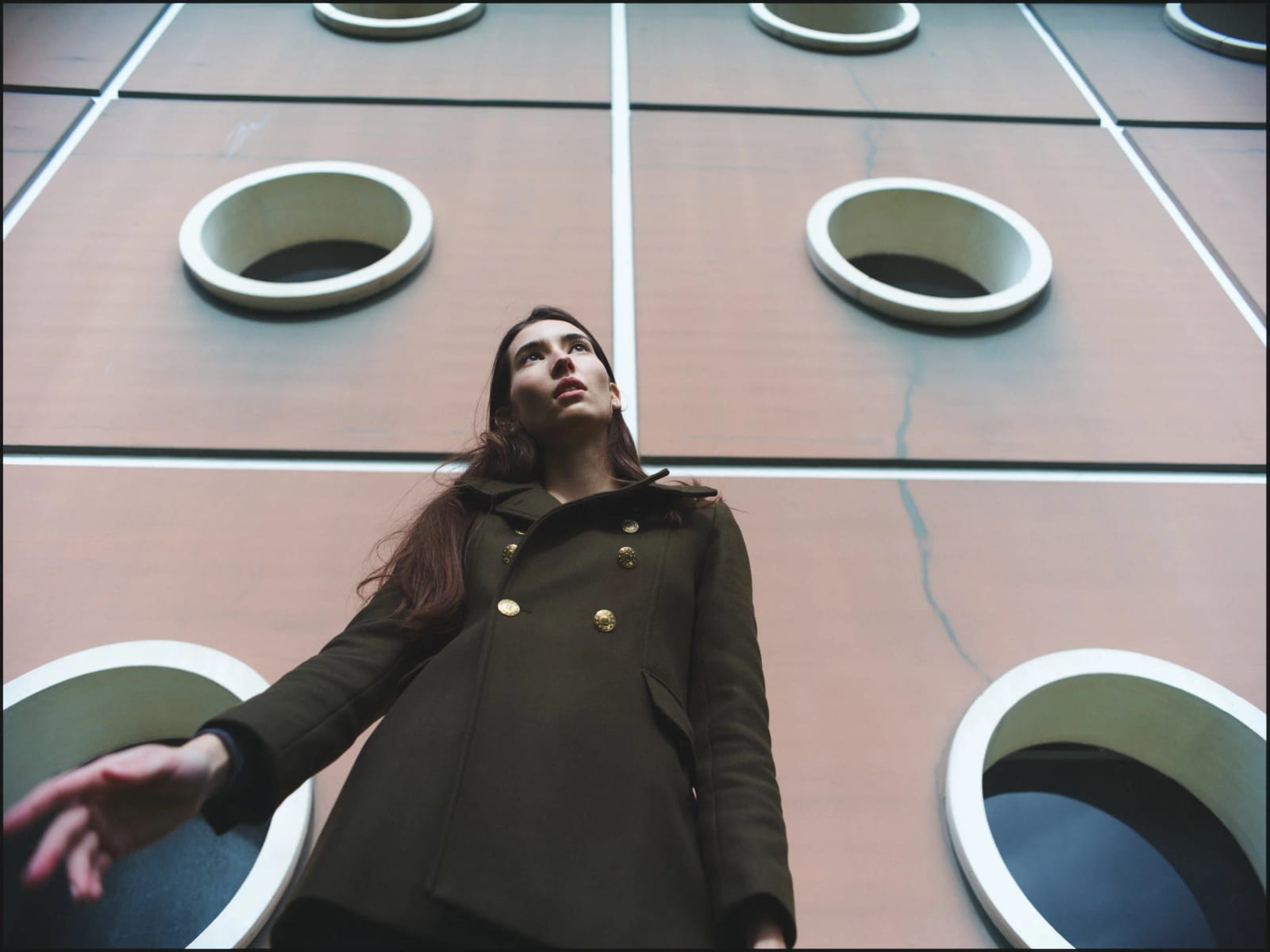 A woman in a dark green coat looking up in front of a wall of round porthole windows.