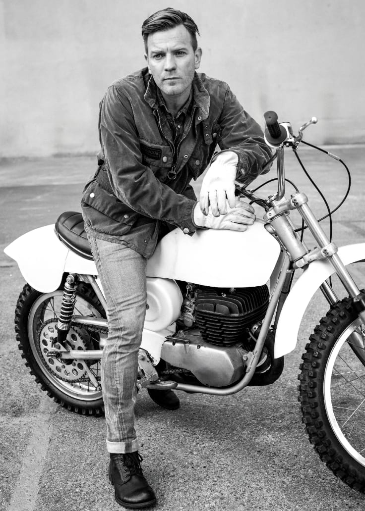 Black and white photograph of Ewan McGregor in a waxed jacket leaning on the handlebars of a motocross bike.