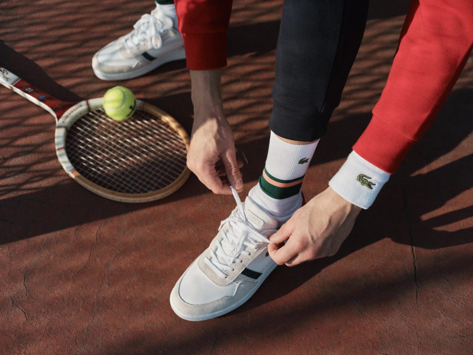A person in red sleeves and white socks tying a white trainer on a clay court, a tennis racket and ball beside them.