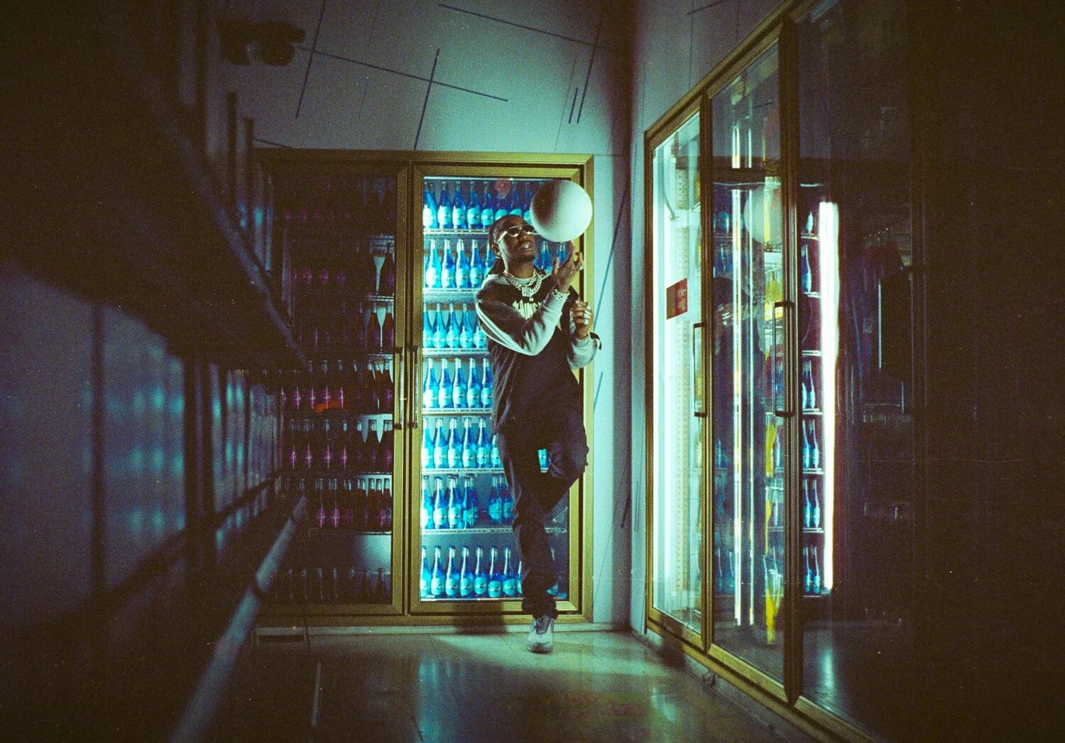 A man standing between glowing drinks fridges in a dark shop, holding a ball up toward a hanging light.