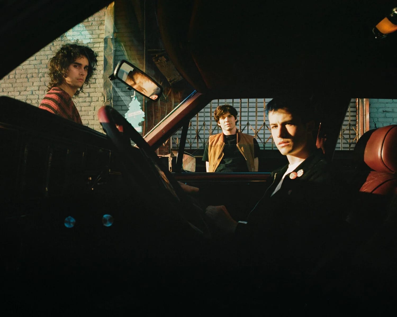 Three young men in a car at night, one in the driver's seat, one leaning in the window and one in the back, lit by a green-lit street.