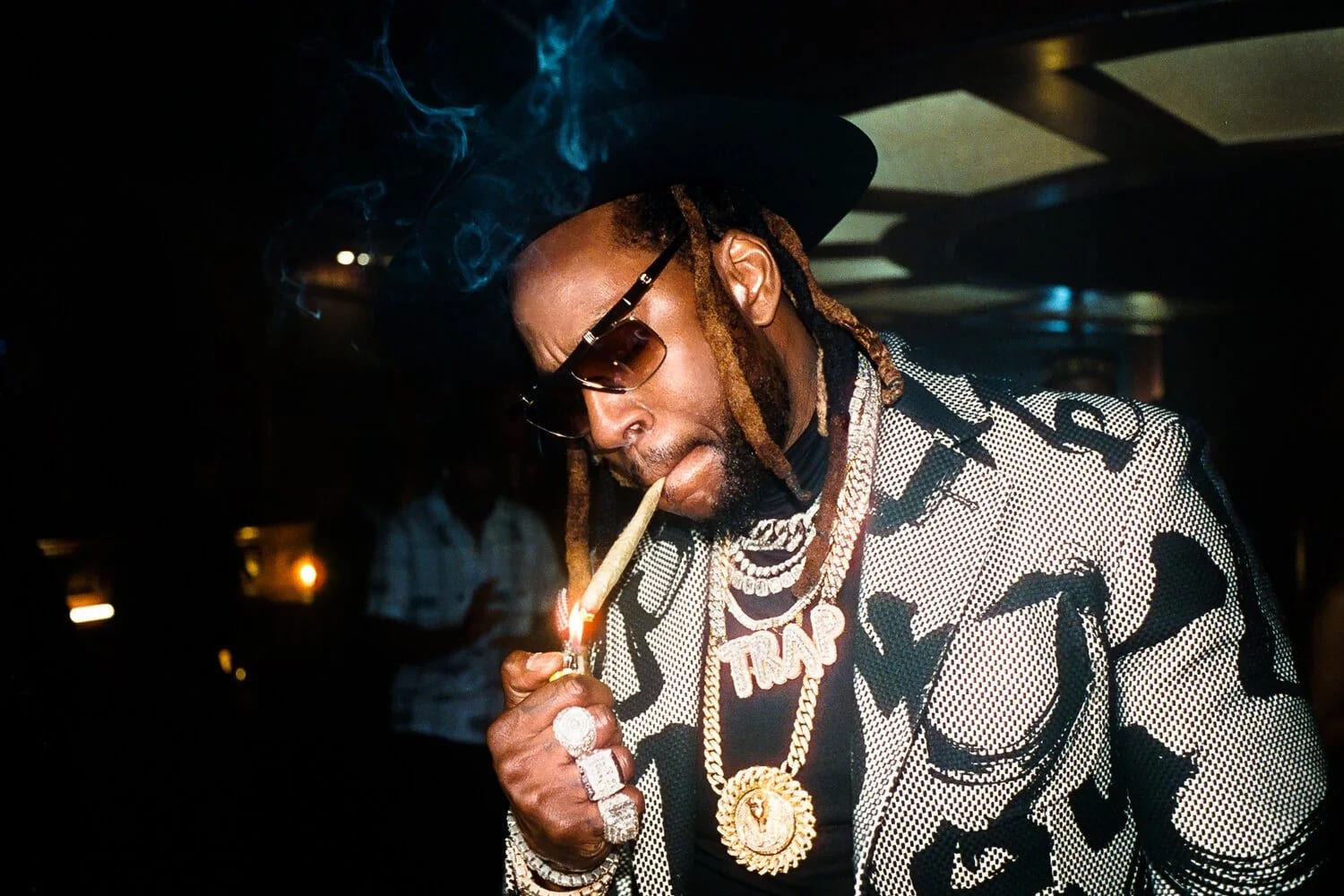 A man in sunglasses and a patterned jacket lighting a cigar, heavy chains around his neck, smoke rising in a dim room.