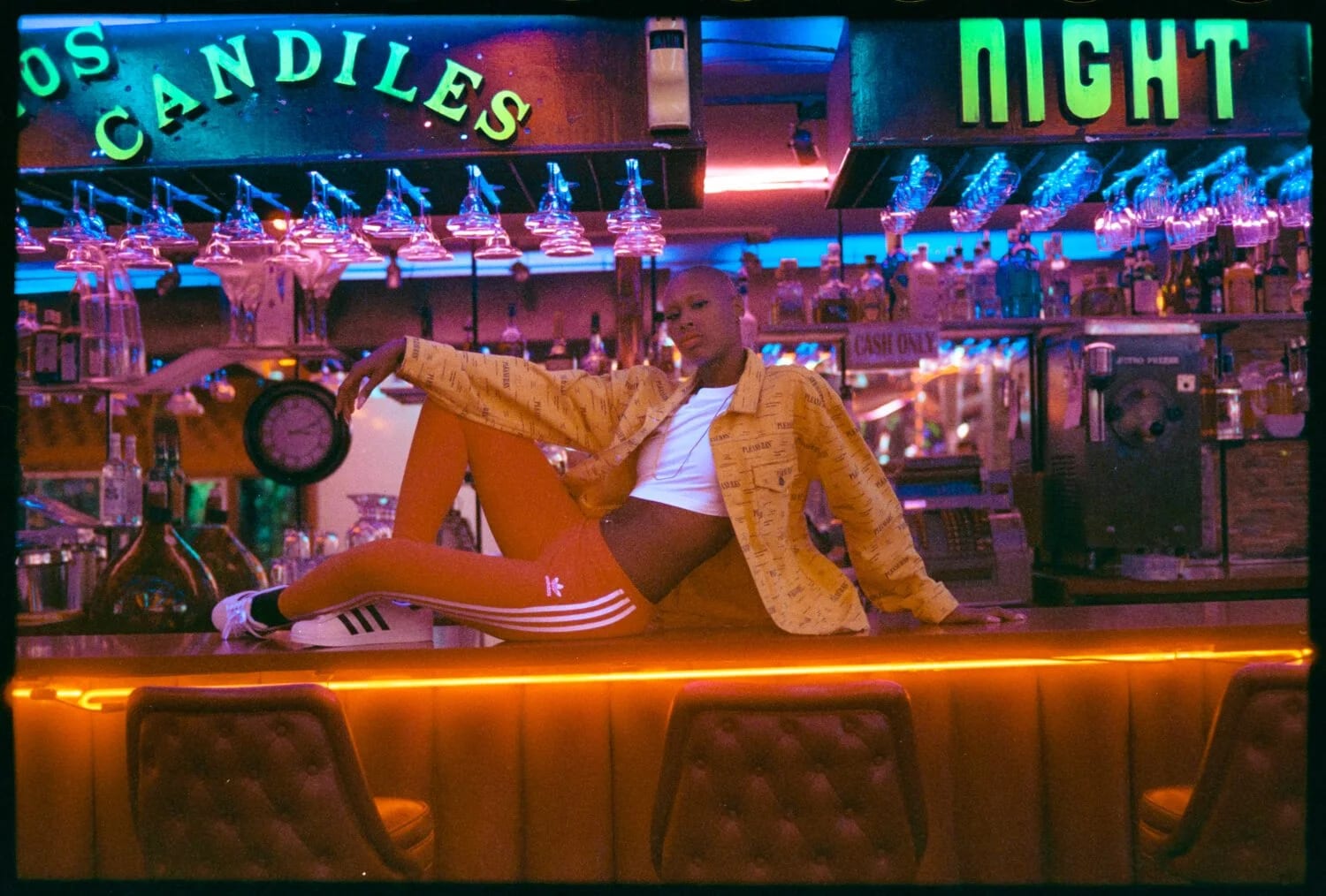 A woman in a yellow jacket and striped adidas trousers reclining along a bar under neon signs reading Los Candiles and Night.