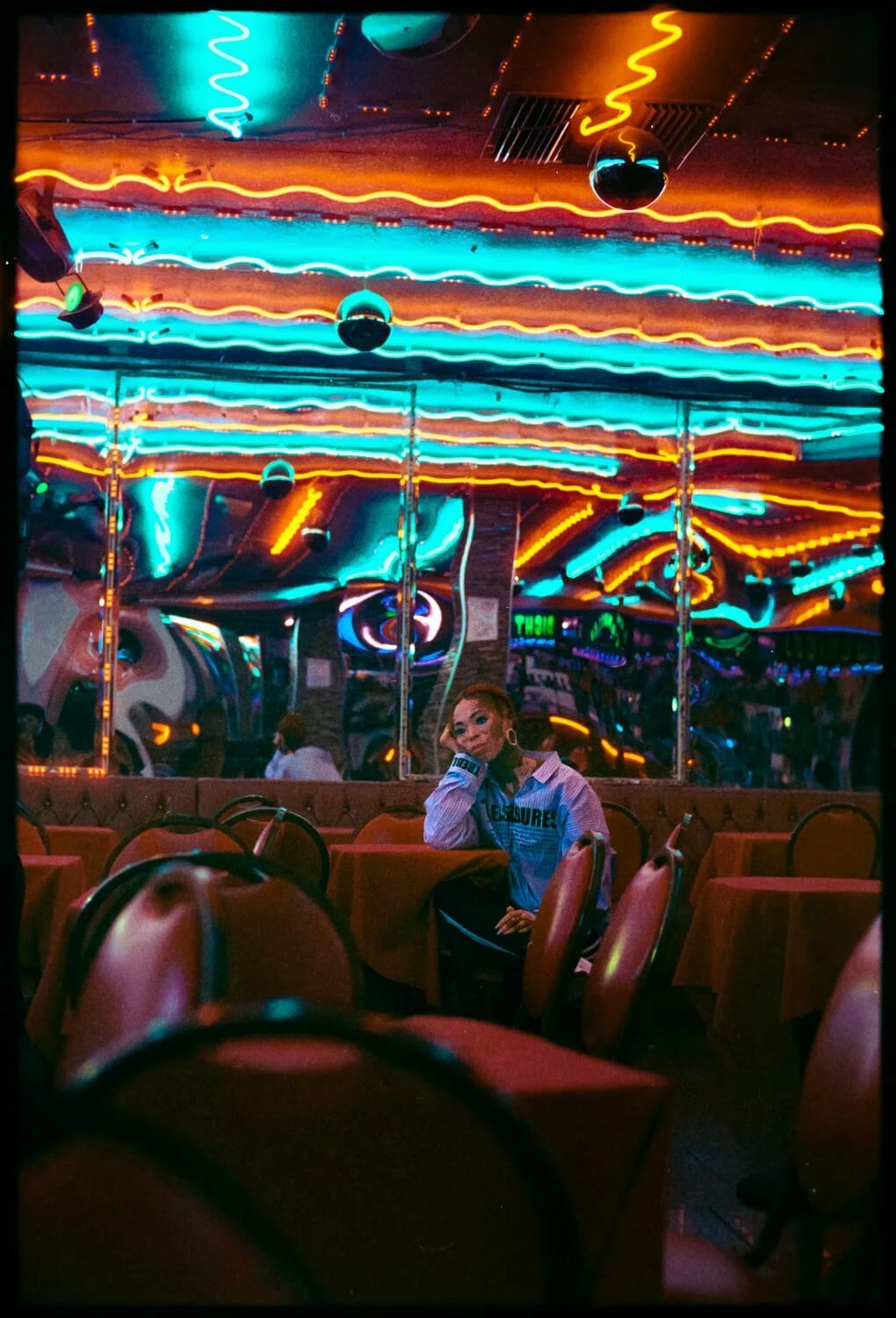 A woman sitting alone in a bar beneath rows of neon tubes in blue, orange and red.