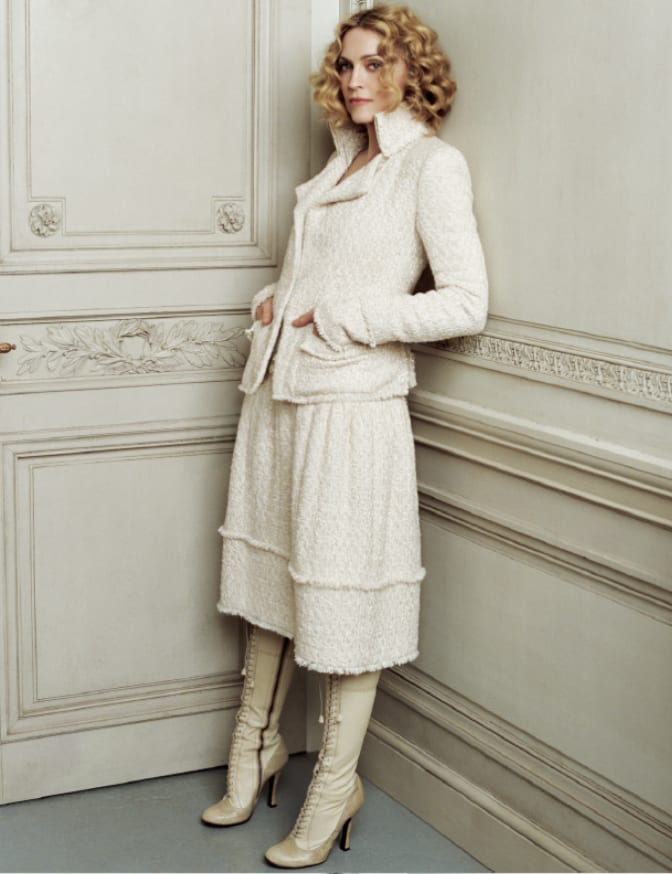A woman with curled blonde hair in a cream tweed jacket and skirt and cream lace-up boots, leaning against an ornate panelled wall.
