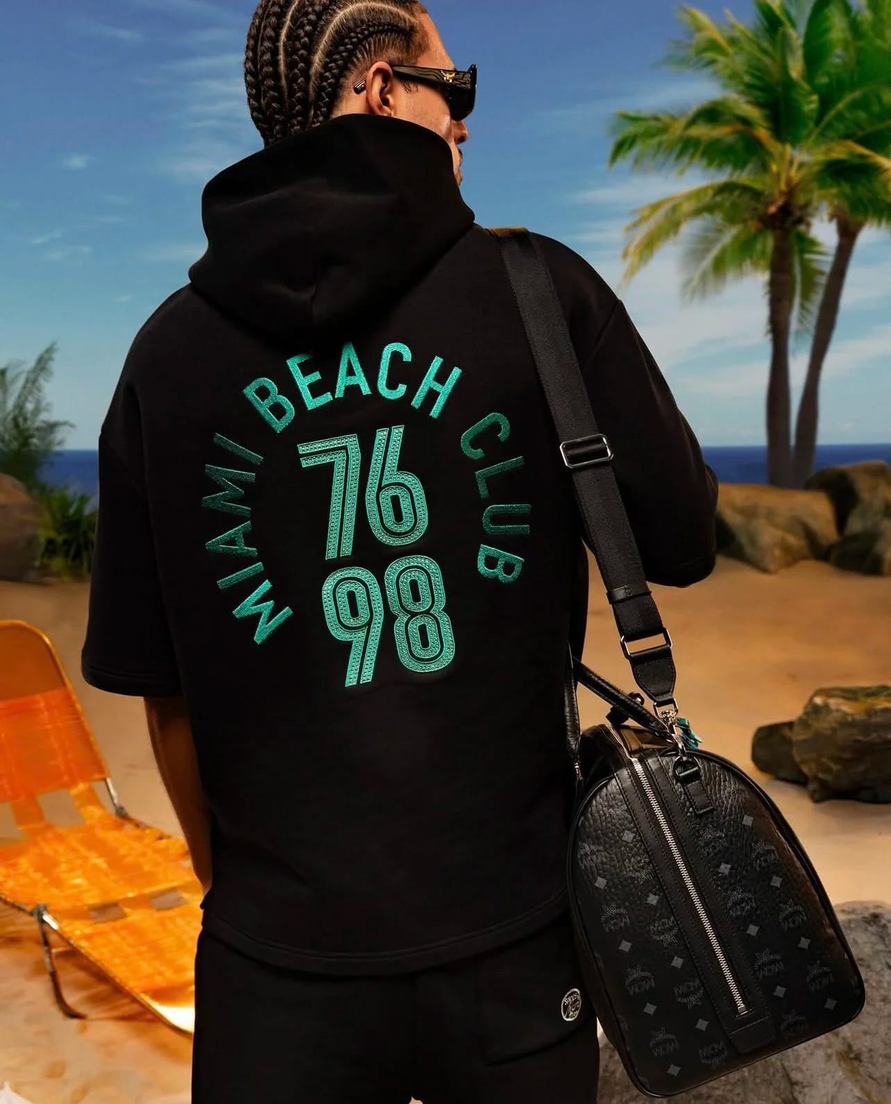 A man with braids seen from behind in a black hoodie reading Miami Beach Club 76 98, carrying a monogrammed bag, palms behind.