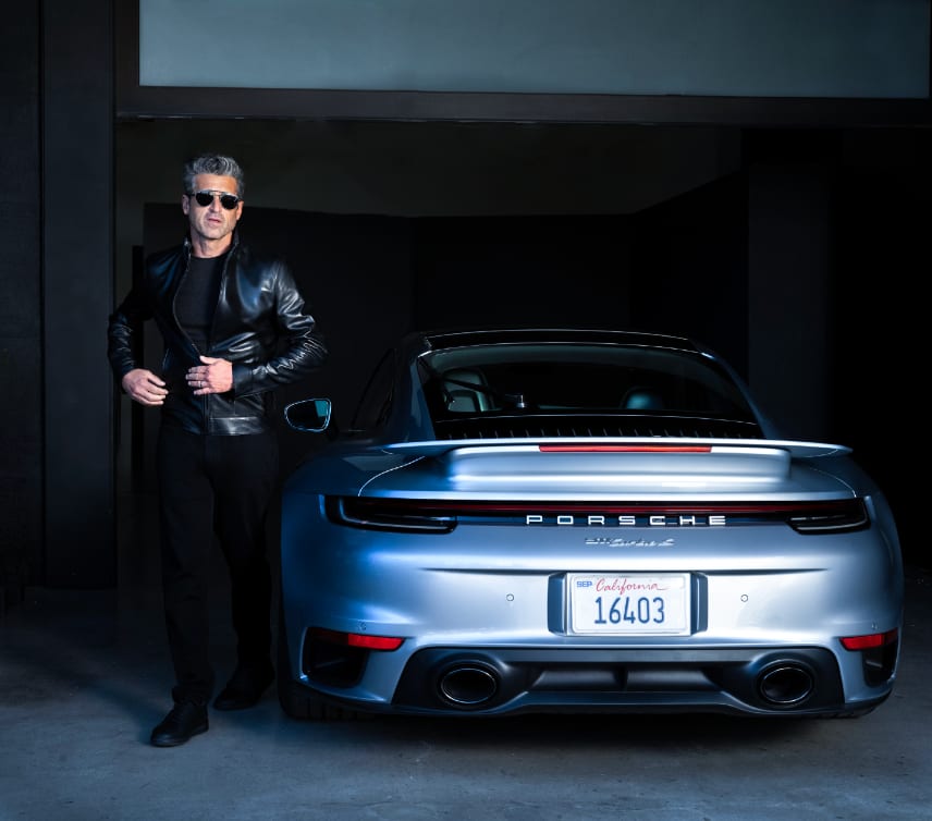 A man in a leather jacket and sunglasses beside a pale blue Porsche parked in a dark garage.