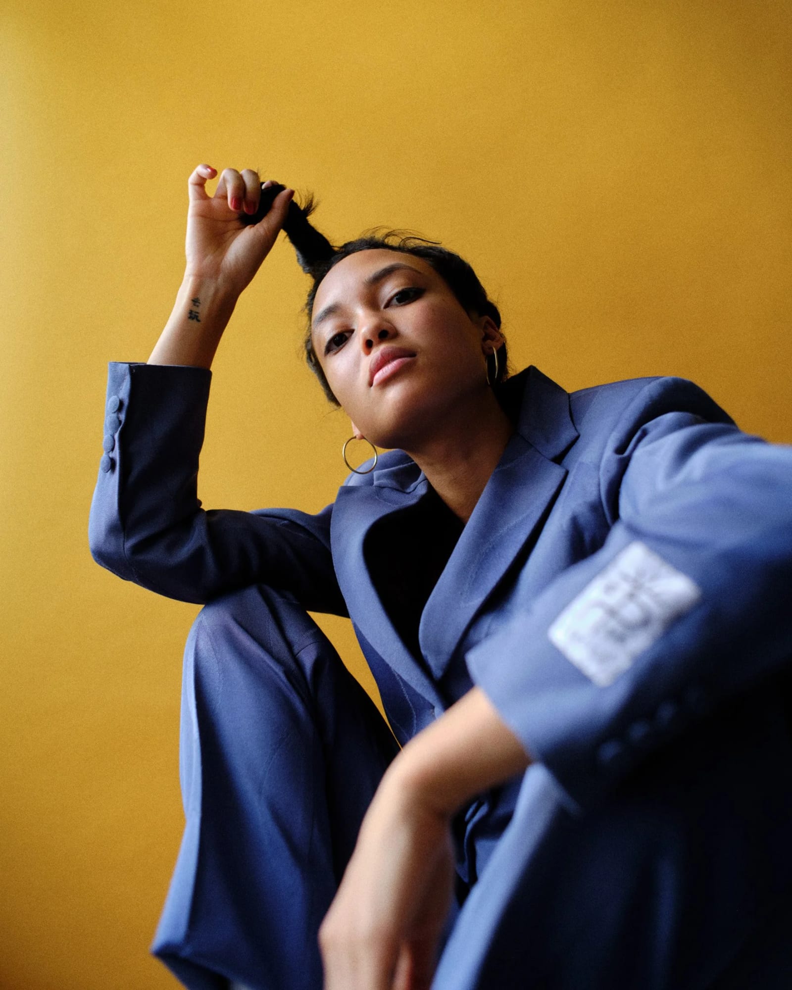 A woman in a blue suit crouching with one hand raised to her hair, looking down at the camera, against a yellow wall.