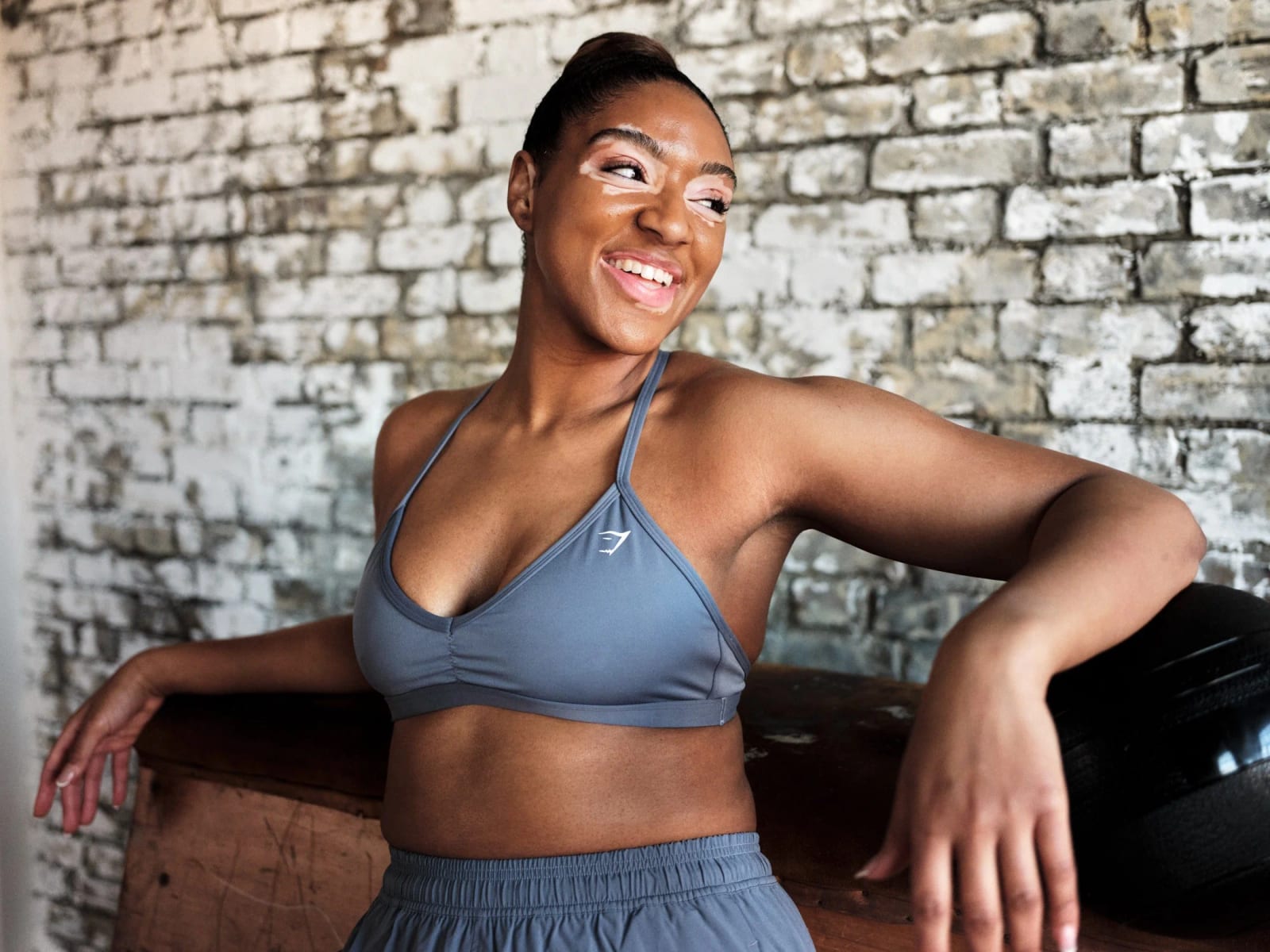 A smiling woman in a grey Gymshark sports bra and shorts leaning against a wooden bench in front of a whitewashed brick wall.