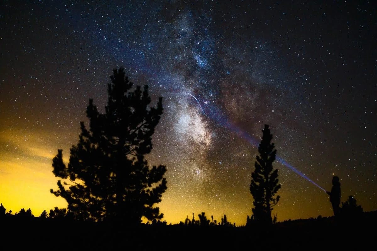 The Milky Way arching over silhouetted pine trees against an orange glow on the horizon.