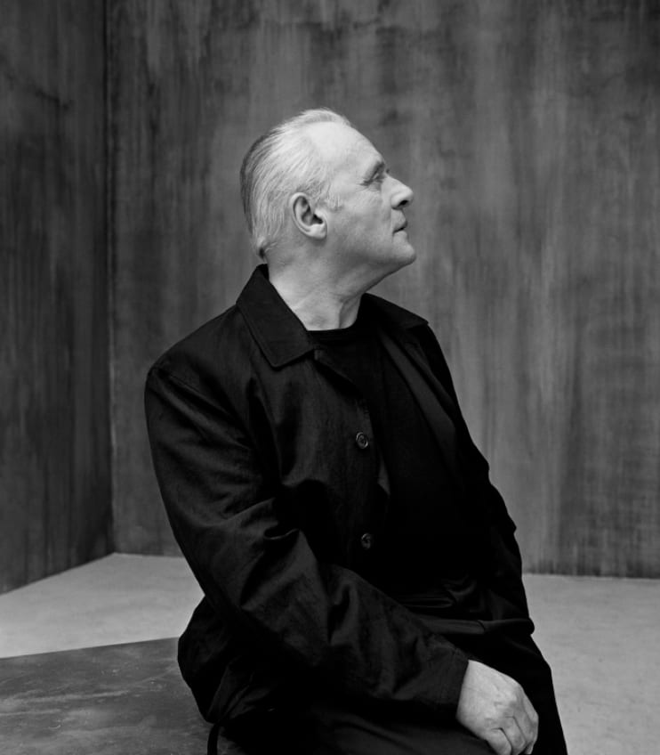 Black and white photograph of a white-haired man in a black shirt seated in a bare room, looking up to the side.