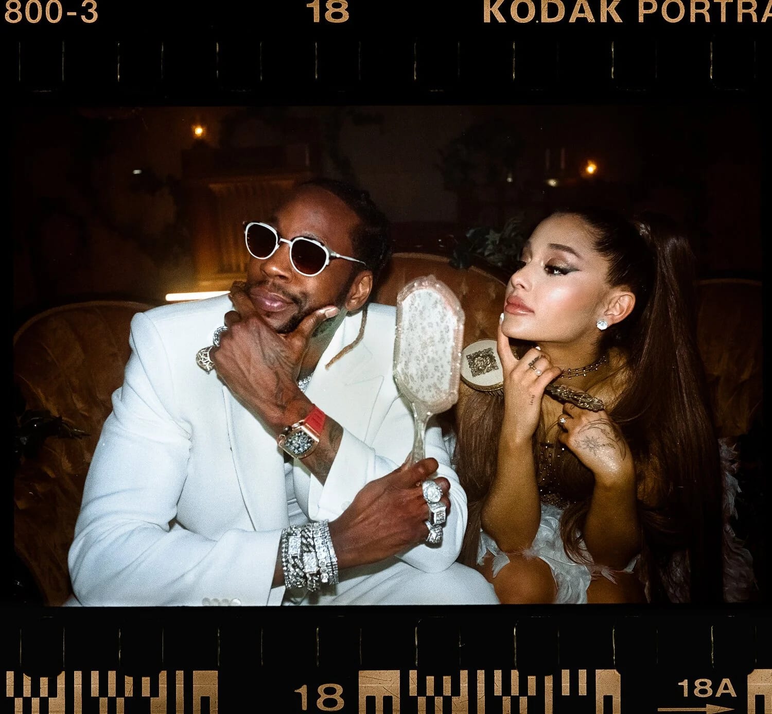 A film-strip frame of a man in a white suit and round sunglasses beside a woman with a high ponytail, both looking at an ornate hand mirror.
