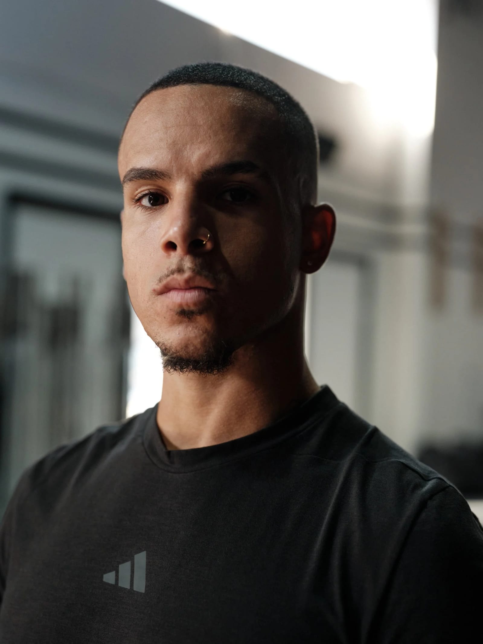 Portrait of a young man with a nose ring in a black adidas T-shirt, looking straight into the camera inside a gym.