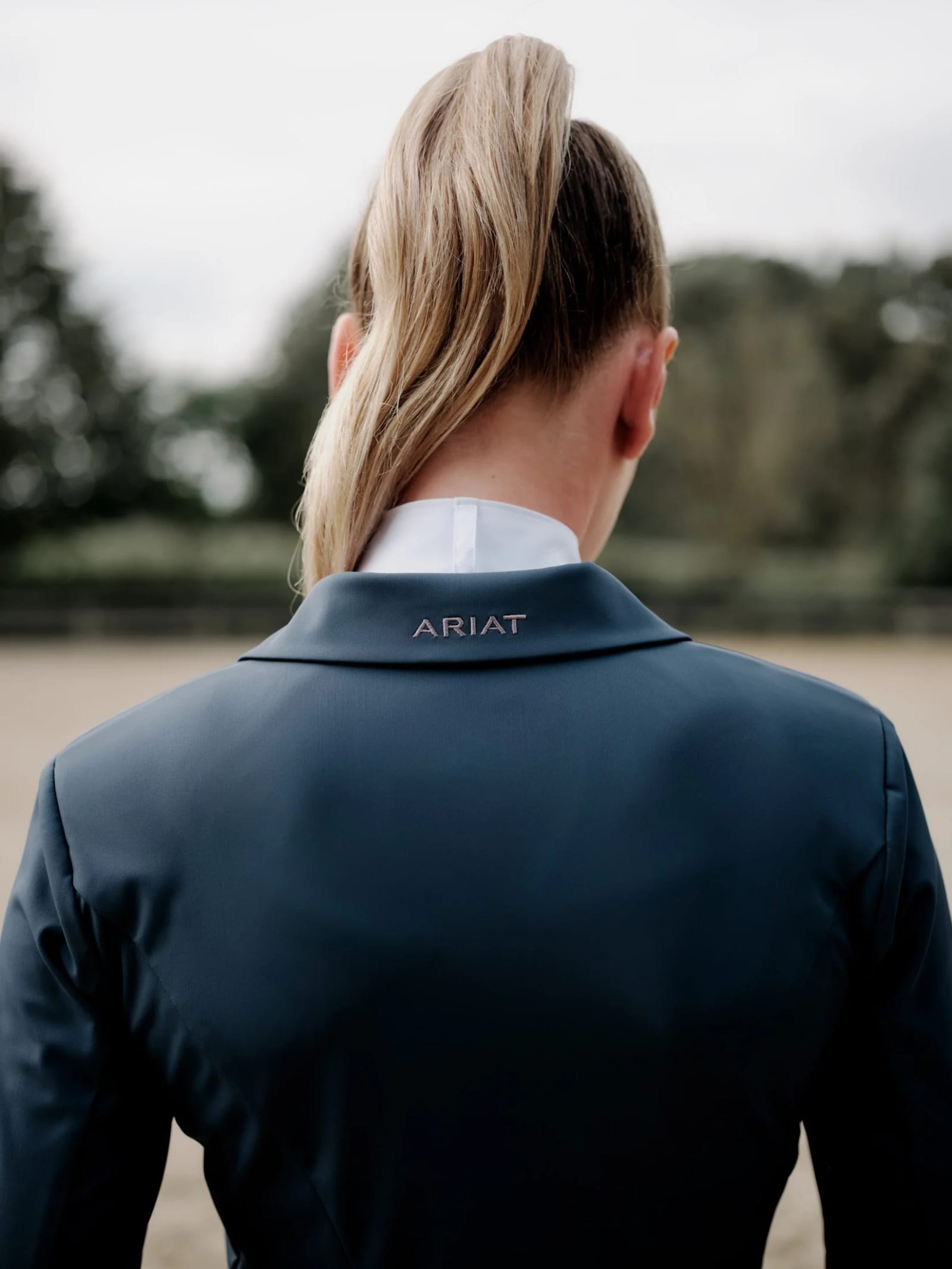 The back of a rider with a blonde ponytail in a navy Ariat show jacket, facing an arena.