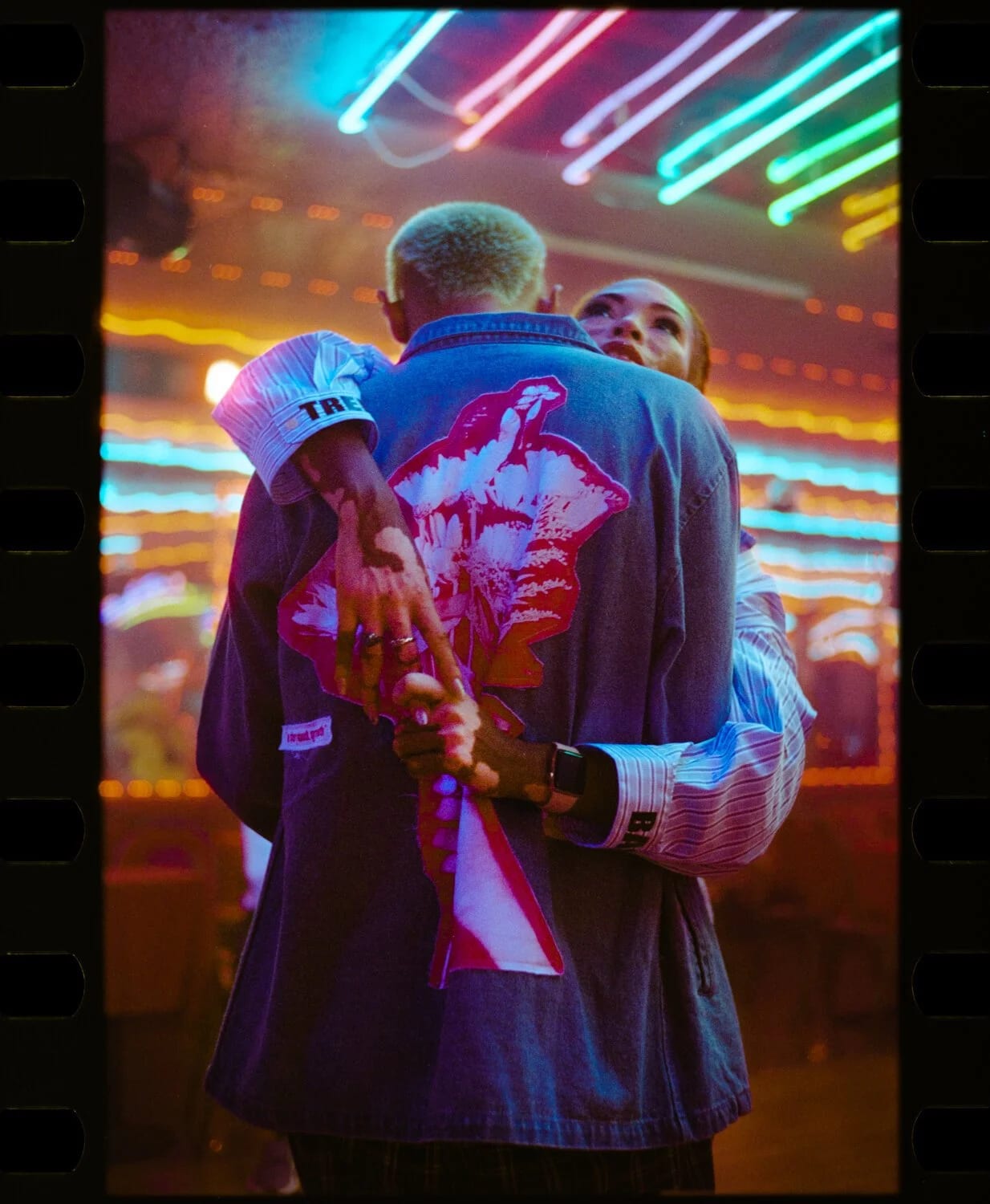 Two people embracing under neon tube lights, one in a denim jacket printed with a pink figure on the back.