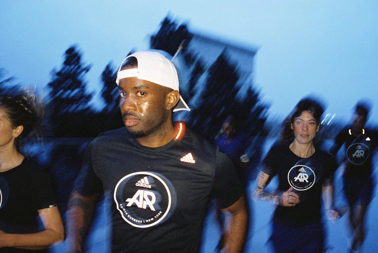 A group of runners in black adidas T-shirts at dusk, the man at the front in a backwards white cap looking at the camera.