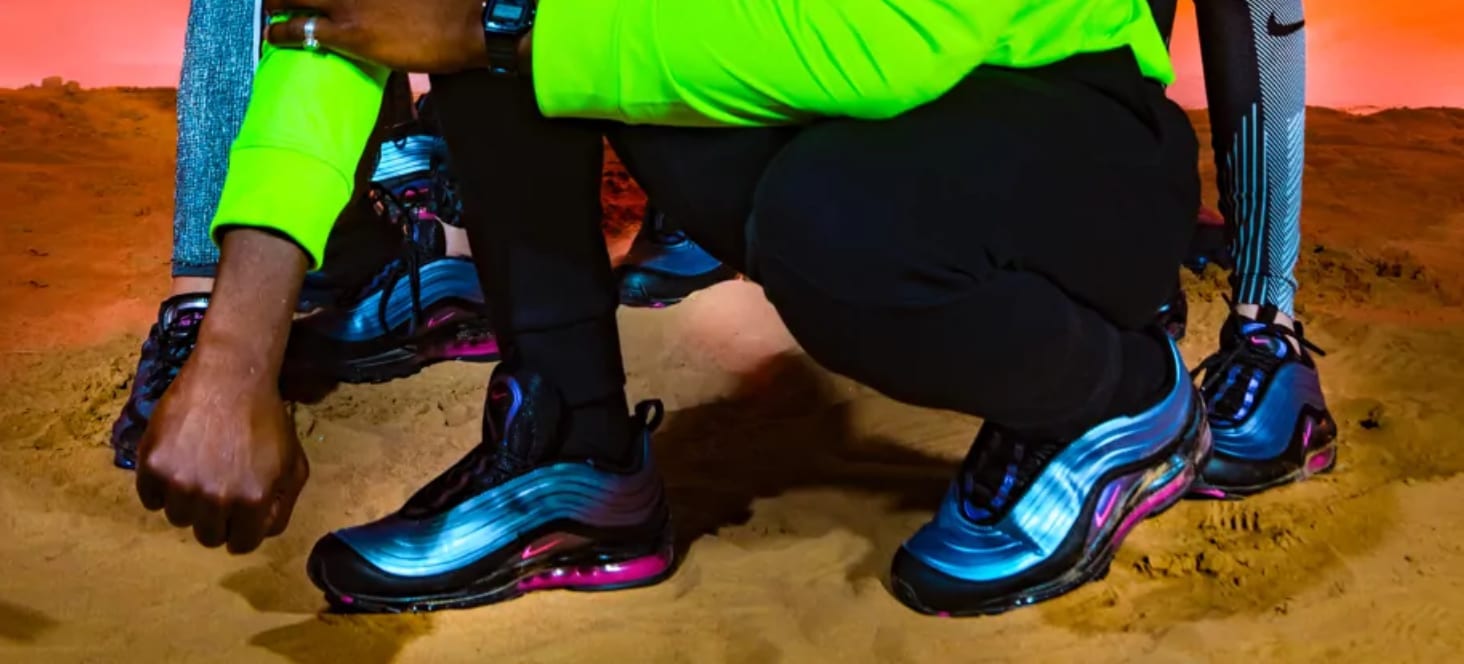 Close-up of blue and pink trainers on sand, a green sleeve and black trousers above.