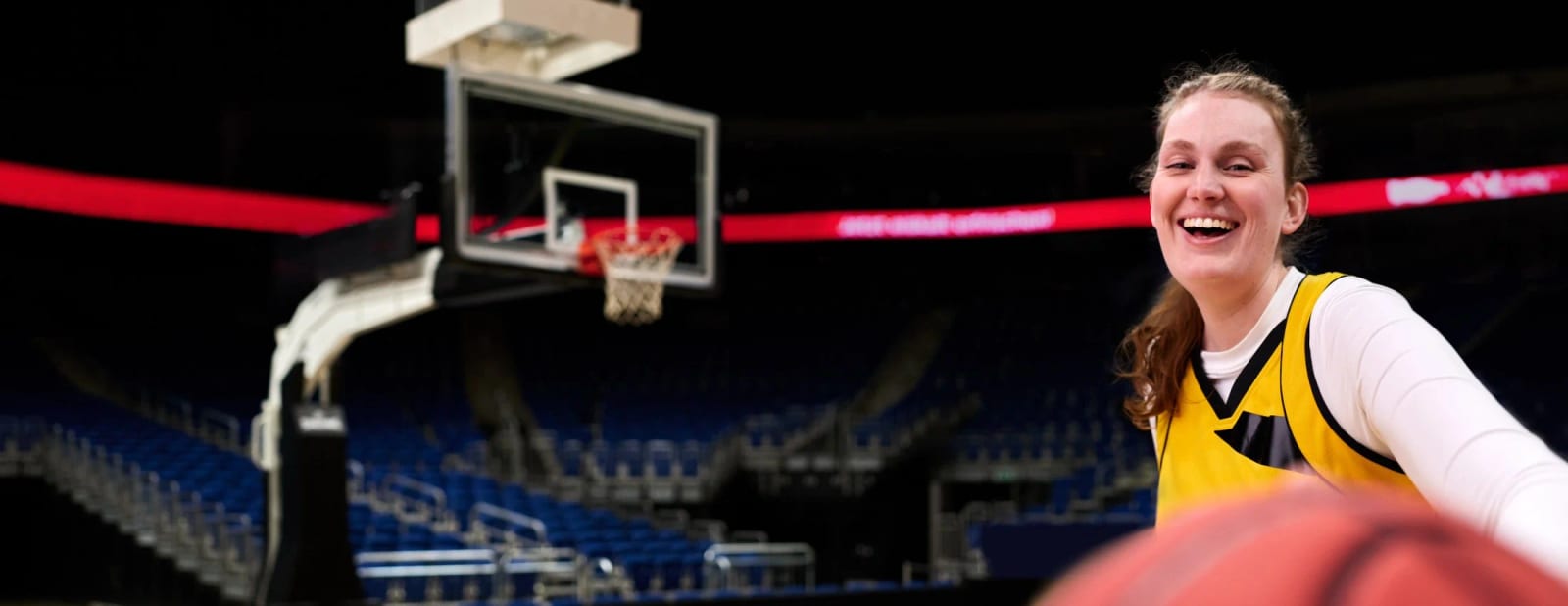 A laughing woman in a yellow basketball jersey beside a hoop in an empty arena, a basketball in the foreground.