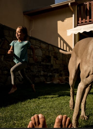 A young girl in a turquoise top running across a lawn beside a large dog, bare feet in the foreground.