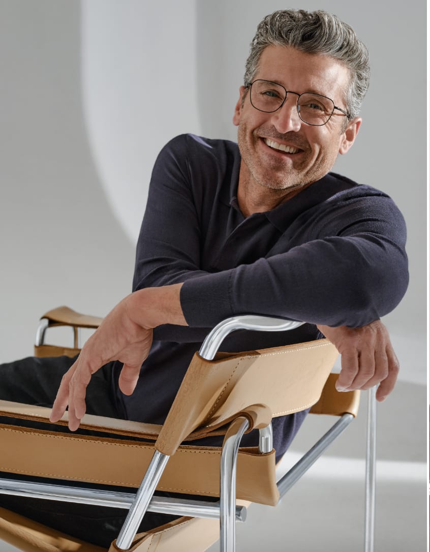 A grey-haired man in glasses and a navy sweater laughing over the back of a tan leather chair.