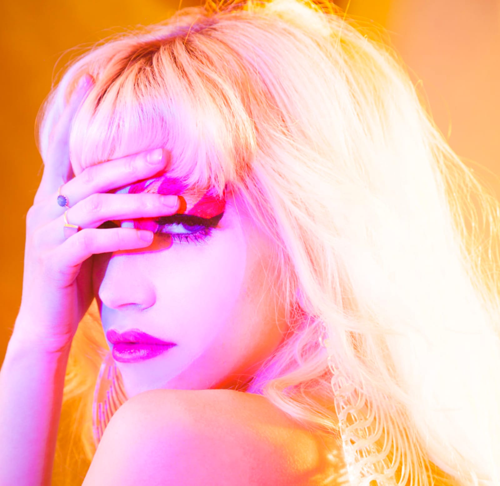 A blonde woman covering one eye with her hand, lit in hot pink and orange.