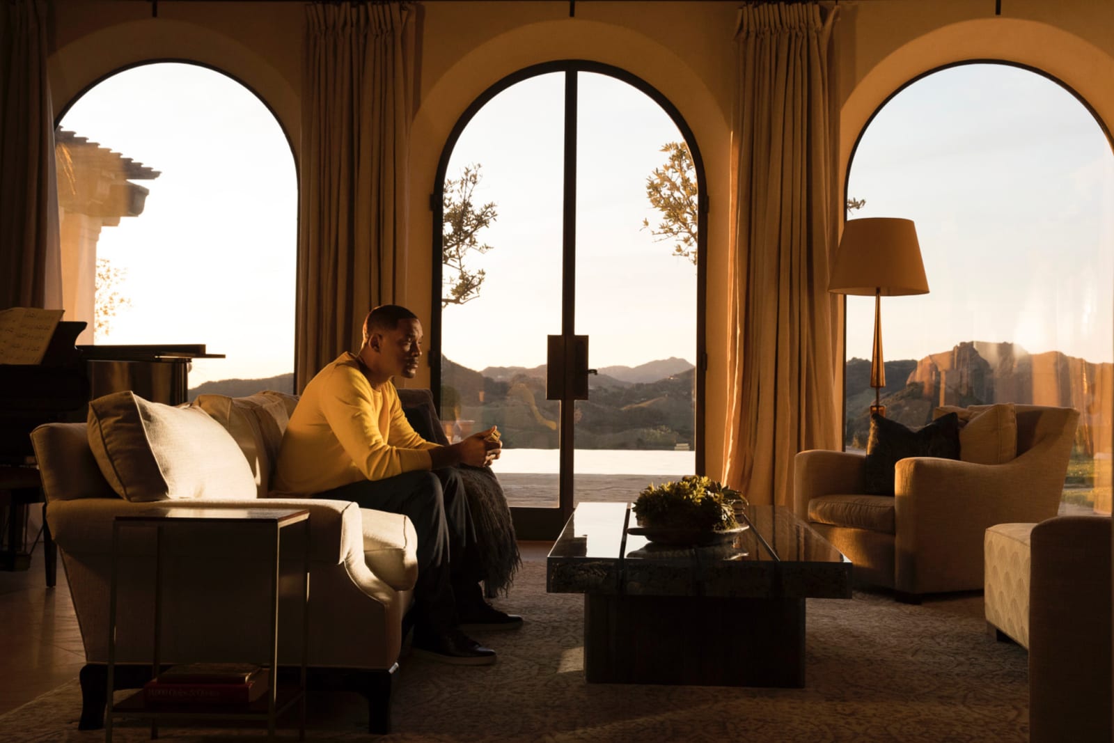 A man in a yellow jumper sitting on a sofa in a warm room, three arched windows opening onto hills at sunset.