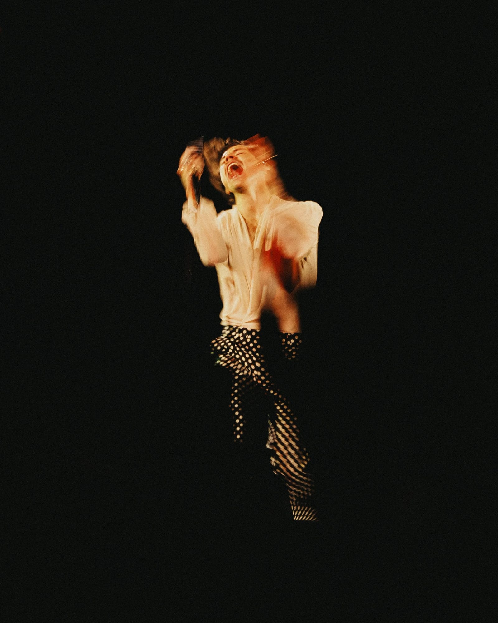 A blurred figure in a loose white shirt and polka-dot trousers mid-motion against black.