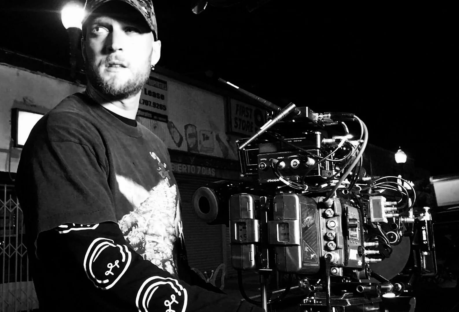Black and white photograph of Colin Tilley in a cap and printed T-shirt beside a cinema camera rig at night.