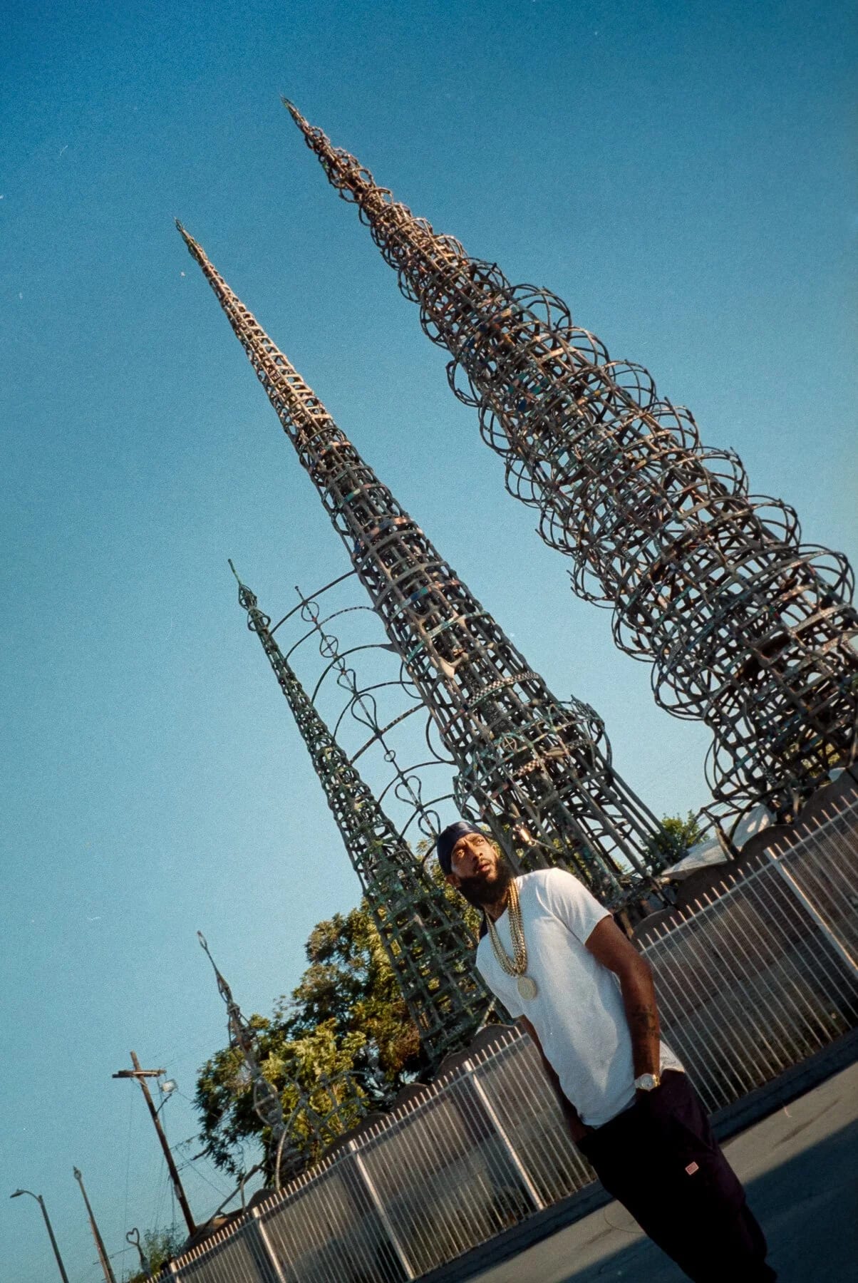 A man in a white T-shirt and gold chain standing in front of tall lattice spires against a blue sky.