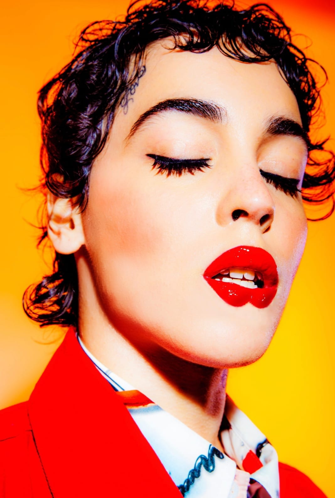 A person with wet curls, red lipstick and long lashes in a red jacket, eyes closed, against orange.