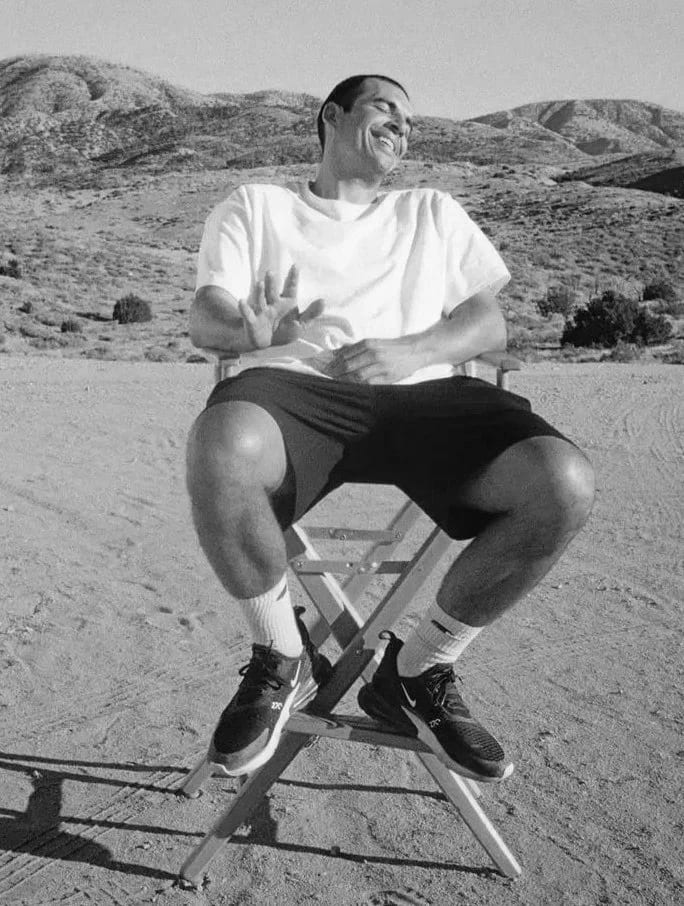 Black and white photograph of Emil Nava in a white T-shirt laughing in a director's chair in a dry hillside landscape.