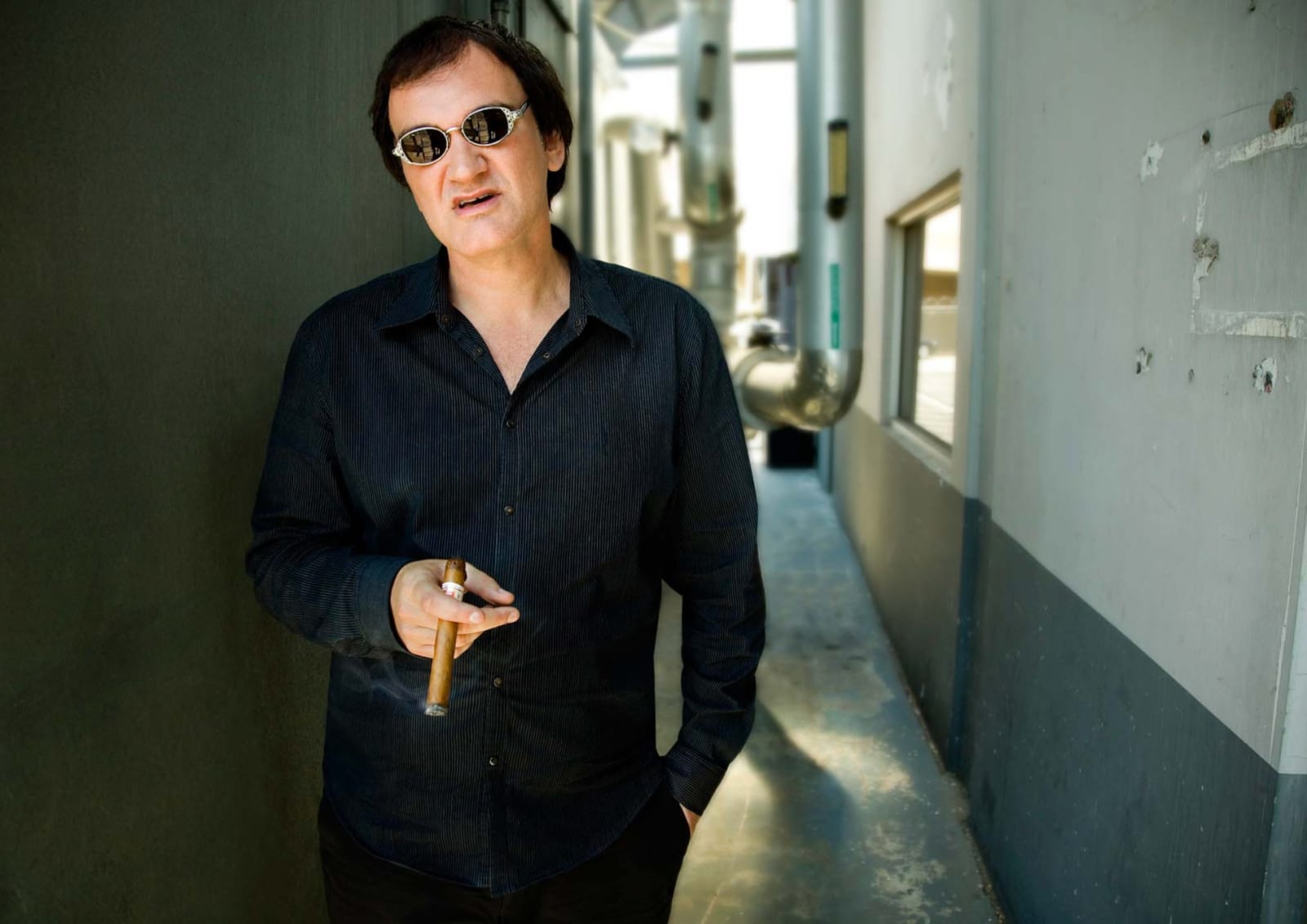 A man in sunglasses and a black shirt holding a cigar in a bare concrete corridor.