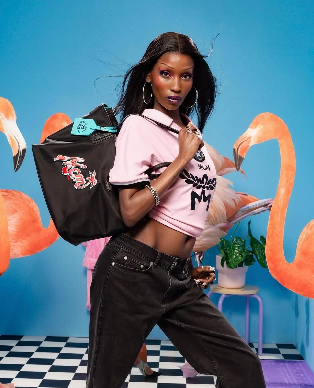 A woman in a pink MCM T-shirt carrying a black bag, posing between two flamingo models on a checkered floor against blue.