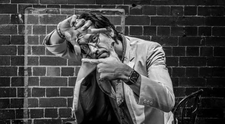 Black and white photograph of Shane Drake in a pale suit framing his face with his hands in front of a brick wall.